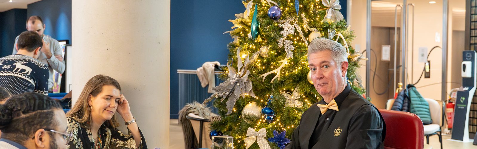 A croupier and guests at a casino table beside a lit Christmas tree at a festive party